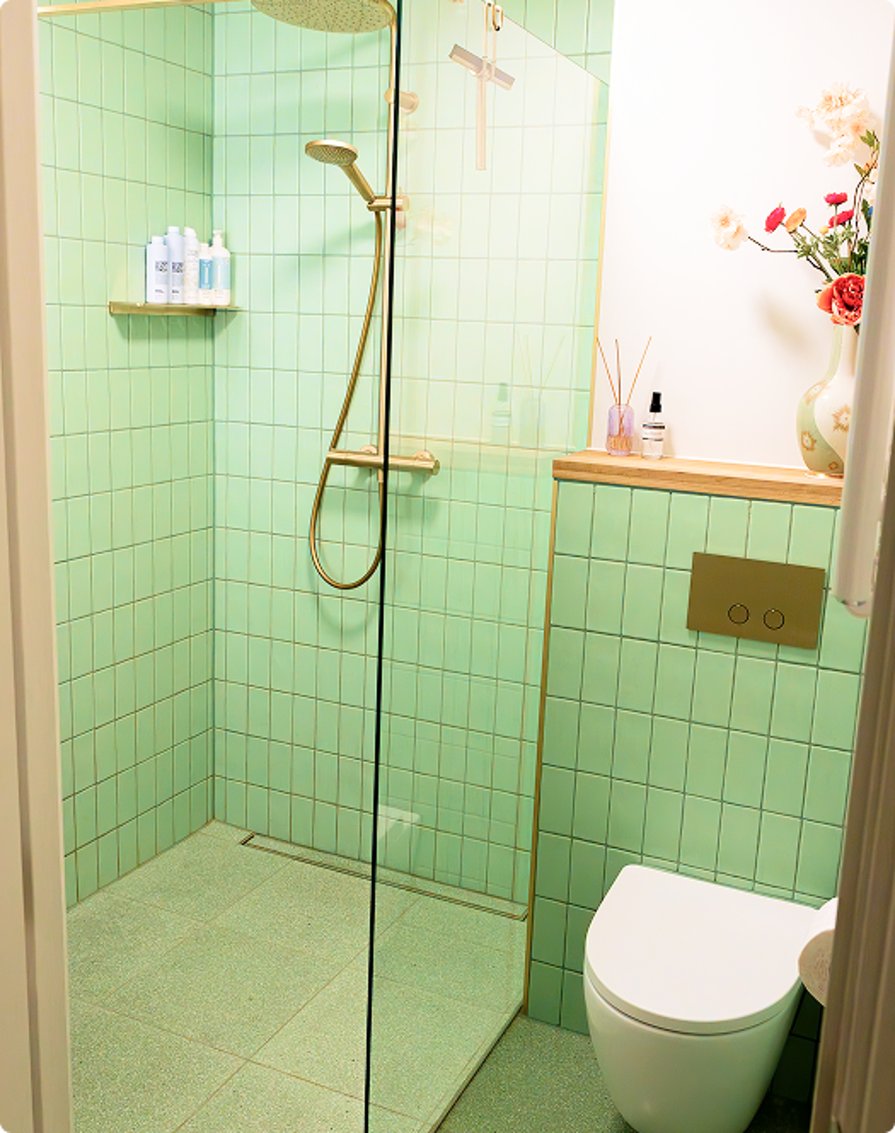 In deze x²o badkamers zie je een moderne inloopdouche met gouden regendouche en handdouche, groene wandtegels, een glazen douchewand, een wandtoilet met gouden bedieningsplaat, en een houten plank met decoratie.