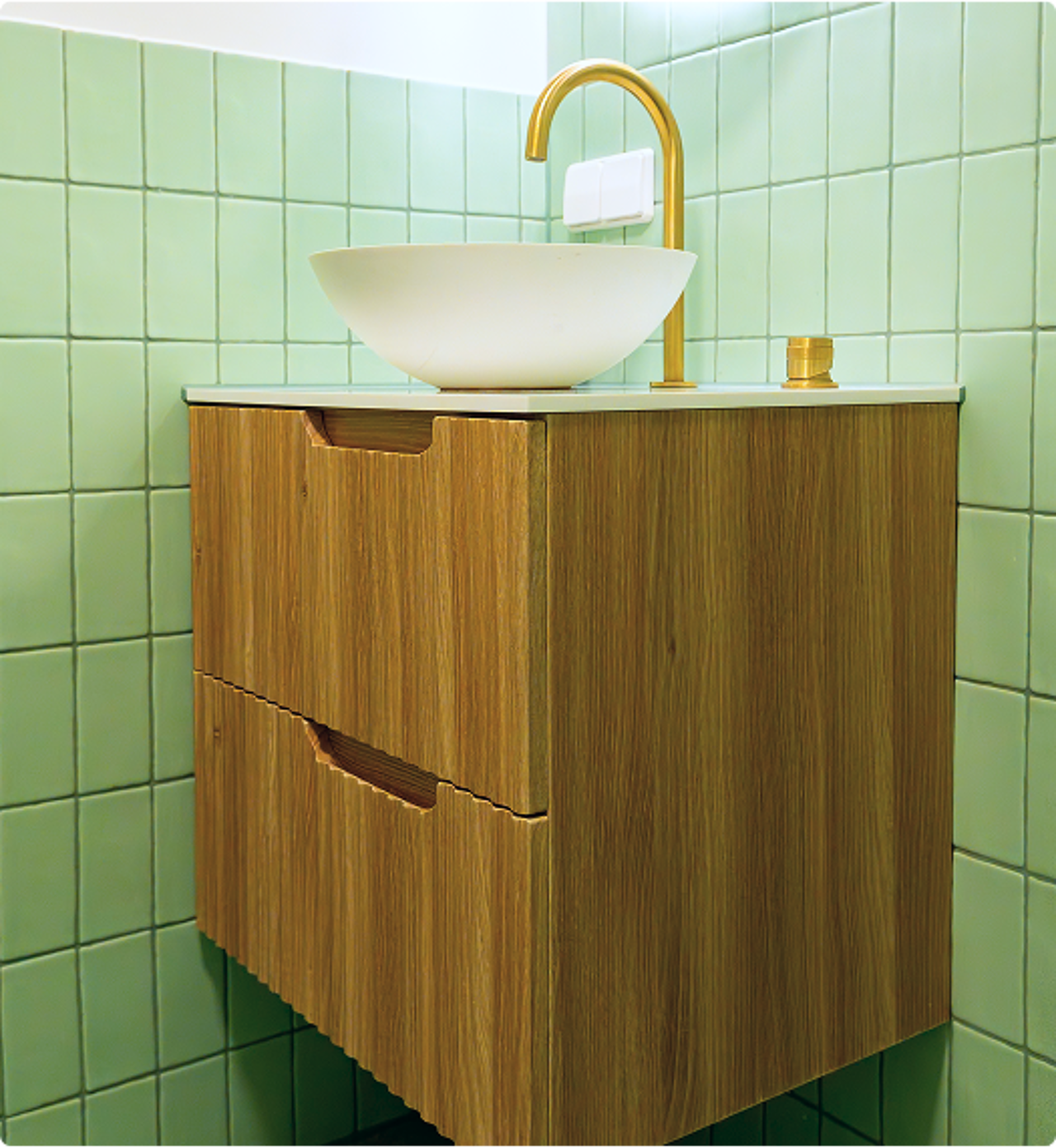 In deze x²o badkamers zie je een houten wastafelmeubel met een witte waskom en een hoge, gouden kraan, geplaatst tegen een wand met mintgroene tegels.