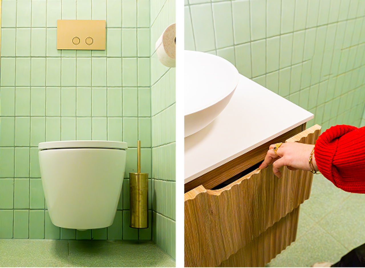  In deze x²o badkamers zie je links een waskom op een houten meubel met gouden kraan, in het midden een wandtoilet met gouden toiletborstel, en rechts een hand die een lade opent van het meubel.