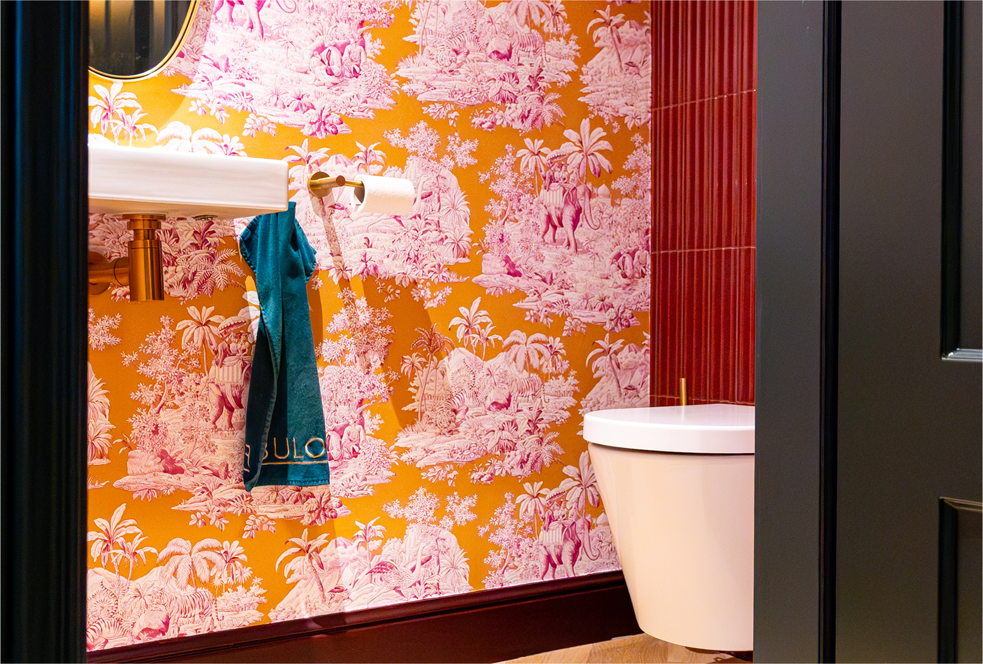 Gouden toiletrolhouder van x²o badkamers met dubbele toiletrollen tegen een rode wandtegel en roze behang.
