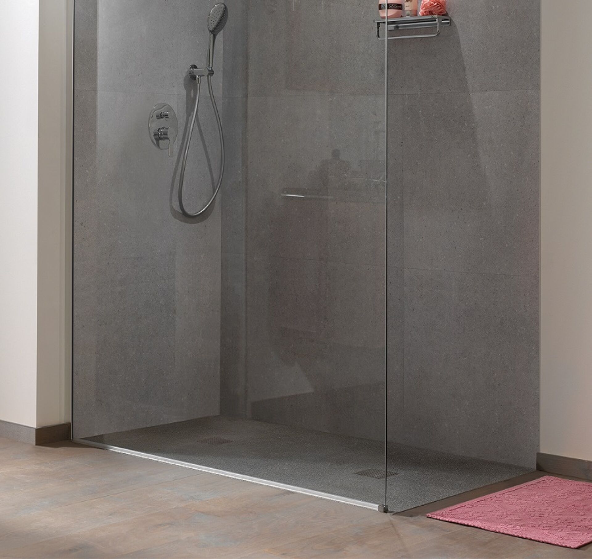 Graue Duschwand mit Glastür, Duschkopf mit Schlauch, Regal mit Produkten an der Wand und rosa Duschmatte auf dem Boden