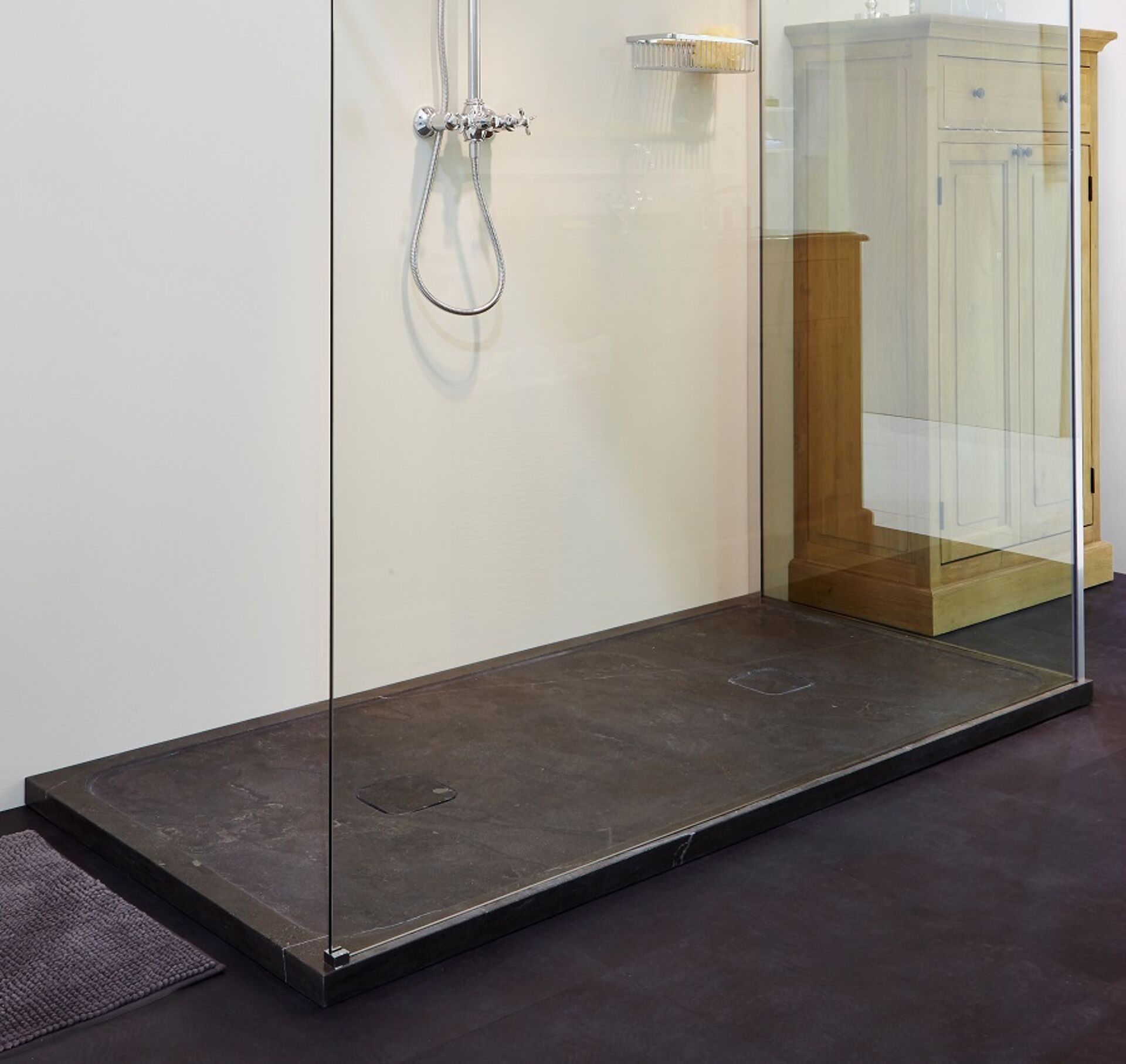 Moderne Dusche mit einem dunklen Steinboden, Glaswand und einem Aufbewahrungskorb für Badezimmerzubehör. Der Boden hat einen minimalistischen Abfluss