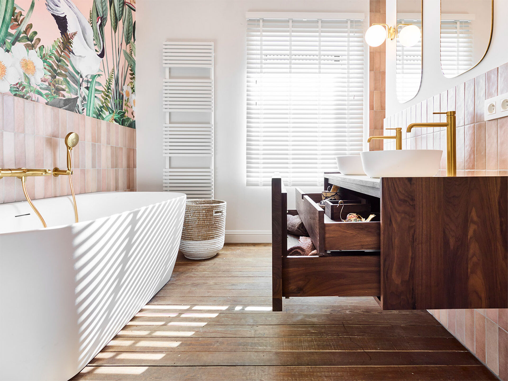 Stilvolles Badezimmer mit einer weißen Badewanne, goldenen Wasserhähnen, tropischer Wanddekoration, einem Holz-Waschbeckenmöbel und einem geöffneten Aufbewahrungsschublade