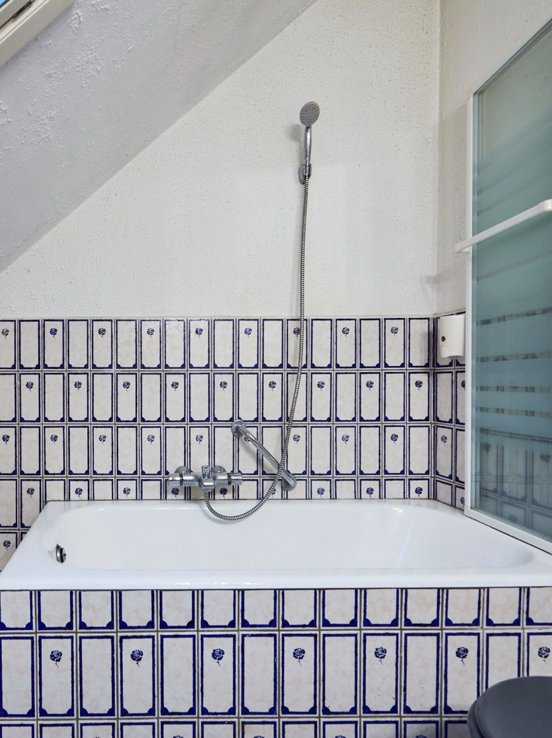 Badewanne mit einer Duschsäule und klassischen Wasserhähnen, umgeben von blauen Fliesenwänden mit Blumenmuster
