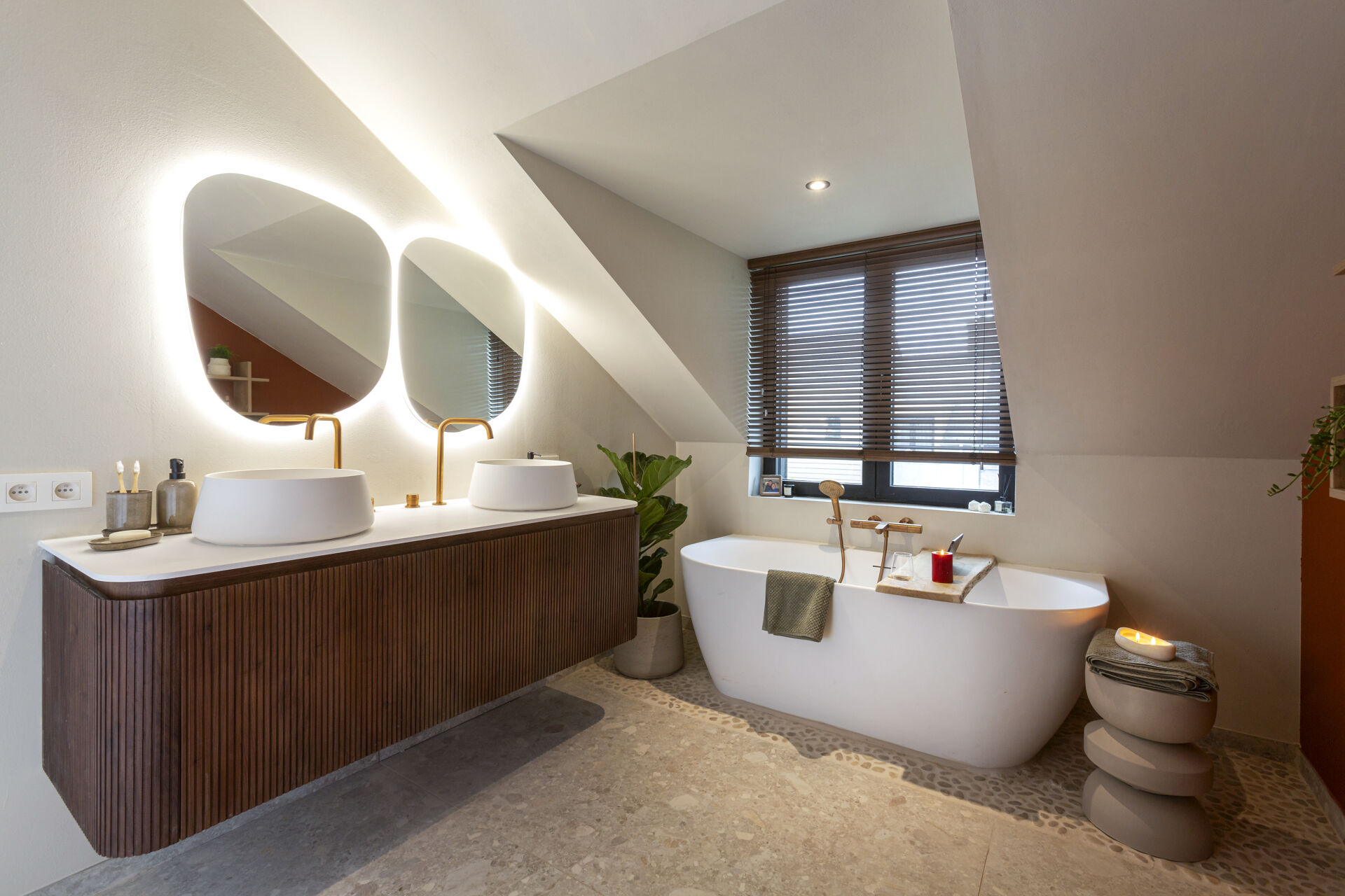 Een stijlvolle x²o badkamers ruimte met een vrijstaand bad, een houten badkamermeubel met twee opbouwkommen en ronde spiegels met geïntegreerde verlichting onder een schuin dak. Er staat een plant naast het bad en een stenen bijzettafel met kaarsjes.