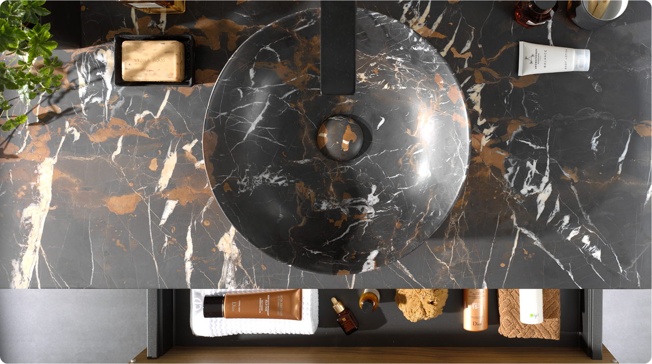 Close-up van een dubbele wastafelopstelling van x²o badkamers met twee ronde waskommen in donker marmerpatroon, zwarte kranen en een houten meubel met verticale groeven. Op het werkblad staan verzorgingsproducten, een tandenborstel en een borstel. Op de achtergrond hangen twee spiegels met geïntegreerde verlichting en daarachter een douchecabine met glazen wand en zwarte accenten.
