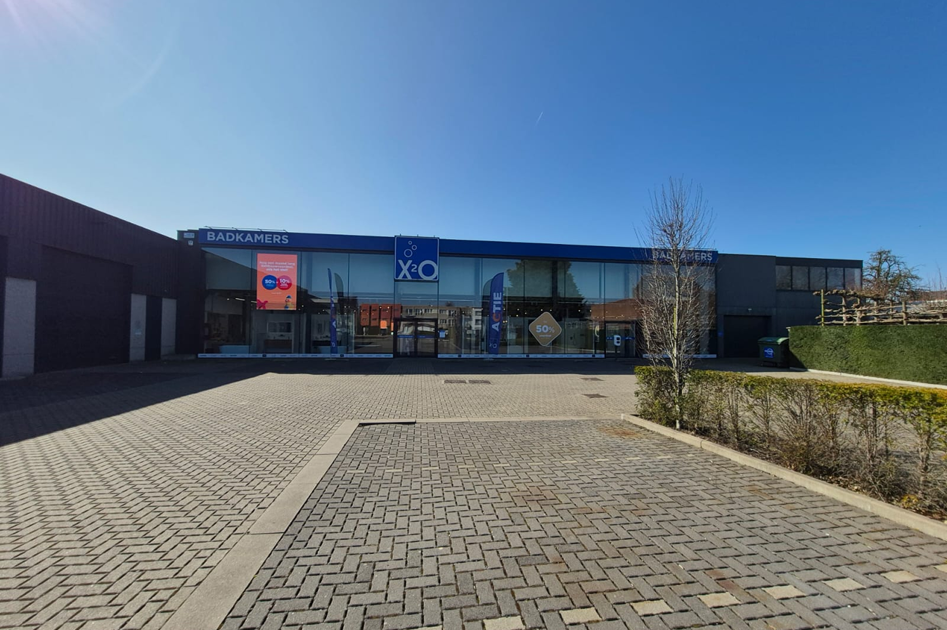 Voorzijde van een XO Badkamers-winkel met een brede glazen gevel, een ruime en lege parkeerplaats en een blauwe lucht op een zonnige dag.