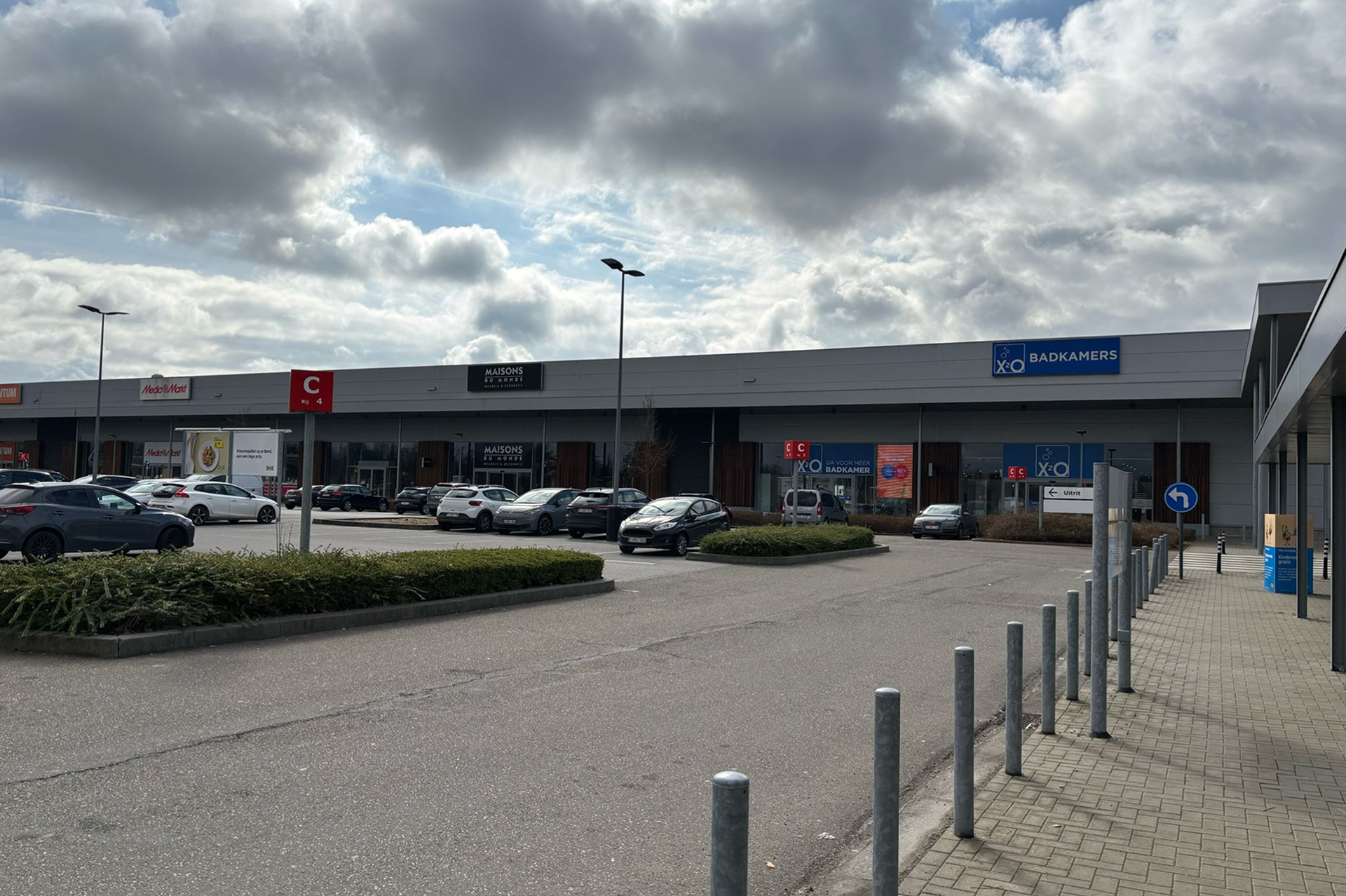 Winkelcentrum met meerdere winkels, waaronder XO Badkamers, met geparkeerde auto's op de voorgrond onder een bewolkte hemel.