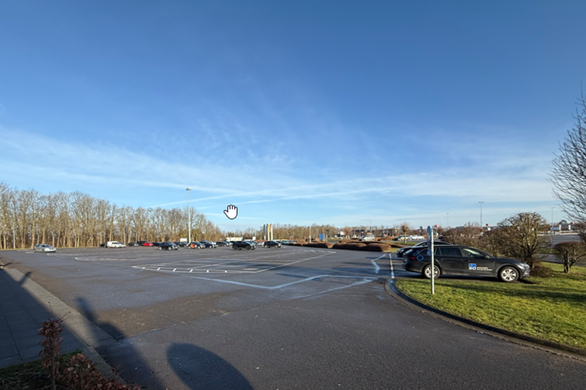 Ruim en bijna leeg parkeerterrein met enkele auto's aan de rand, onder een heldere blauwe lucht met bomen op de achtergrond.