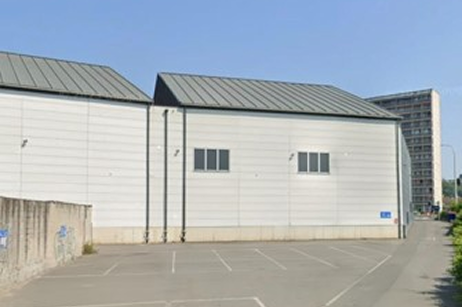 Zijaanzicht van een wit industrieel gebouw met een metalen dak, enkele ramen en lege parkeerplaatsen op een zonnige dag.