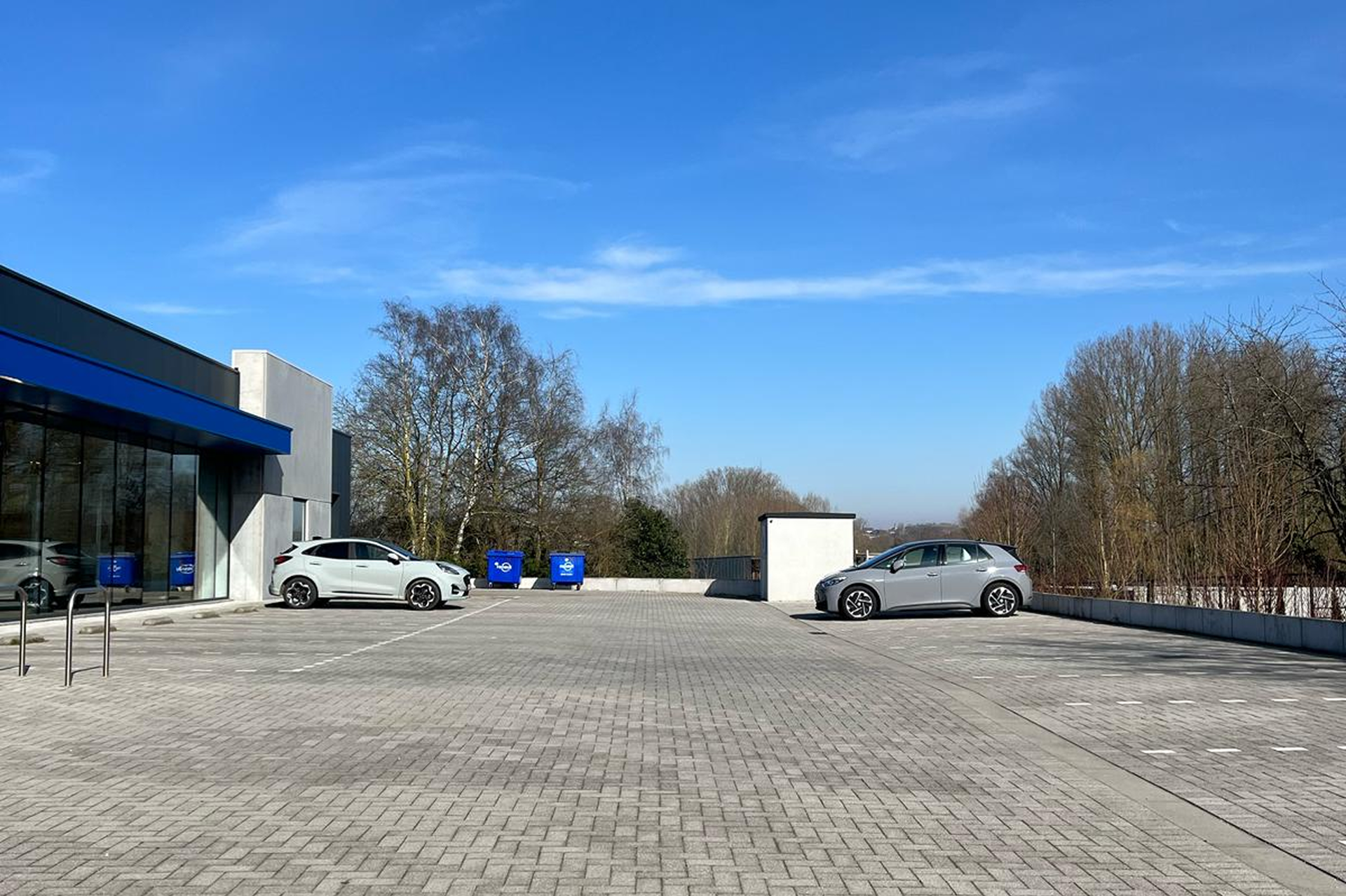 Ruim en bijna leeg parkeerterrein met enkele auto's en blauwe afvalcontainers, gelegen naast een gebouw met glazen gevel onder een heldere blauwe lucht.