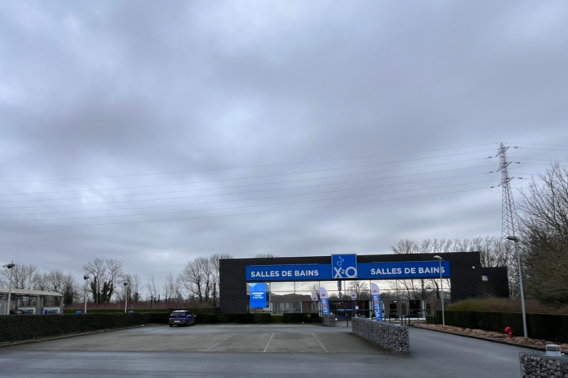 Ingang van een X2O Badkamers-winkel met dubbele gevelbelettering “Salles de Bains”, een grotendeels lege parkeerplaats en een bewolkte lucht.